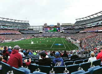 A large sports stadium filled with spectators. The field is green with white markings, and players in red and white uniforms are actively engaged in a game. The stands are packed with people wearing various colored clothing, and some sections of the crowd are more densely populated. Digital screens and advertisements are visible around the stadium, adding to the vibrant atmosphere.