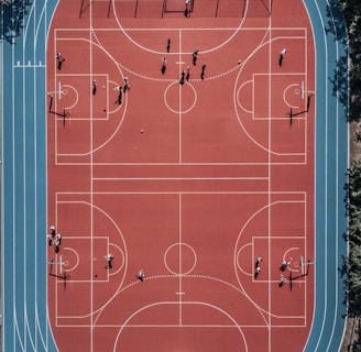 An aerial view of a large, vibrant sports complex featuring a red basketball court with multiple hoops and a surrounding blue running track. Several people are scattered across the area, engaged in various activities.