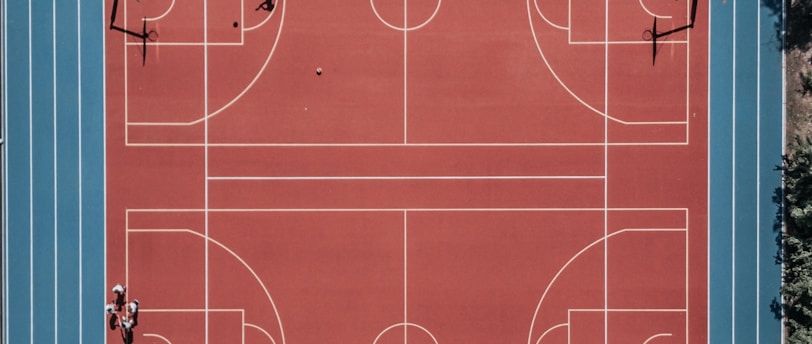 An aerial view of a large, vibrant sports complex featuring a red basketball court with multiple hoops and a surrounding blue running track. Several people are scattered across the area, engaged in various activities.