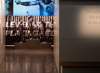 A dimly lit display features statues of athletes standing on a podium. They are positioned in front of a backdrop with the words 'LEVELING THE PLAYING FIELD' in bold letters. On the wall to the right, a softly lit quote is partially visible. The overall ambiance suggests a museum or exhibition setting.