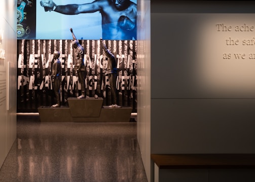 A dimly lit display features statues of athletes standing on a podium. They are positioned in front of a backdrop with the words 'LEVELING THE PLAYING FIELD' in bold letters. On the wall to the right, a softly lit quote is partially visible. The overall ambiance suggests a museum or exhibition setting.