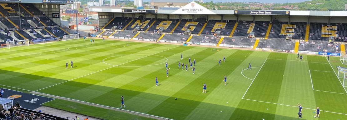 A sports stadium with a well-maintained grass field is depicted. Players are spread across the field, engaging in warm-up activities. The stands are partially filled with spectators, and the stadium is surrounded by urban buildings under a partly cloudy sky.
