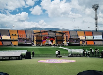 A sports stadium with a large stage set up on the field, hosting an outdoor event. Spectators wearing colorful hats sit in the foreground. The seating areas display vibrant colors, and a clear blue sky with scattered clouds is visible. Large screens display graphics related to the event.