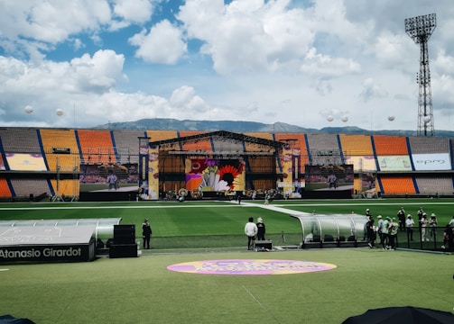 A sports stadium with a large stage set up on the field, hosting an outdoor event. Spectators wearing colorful hats sit in the foreground. The seating areas display vibrant colors, and a clear blue sky with scattered clouds is visible. Large screens display graphics related to the event.